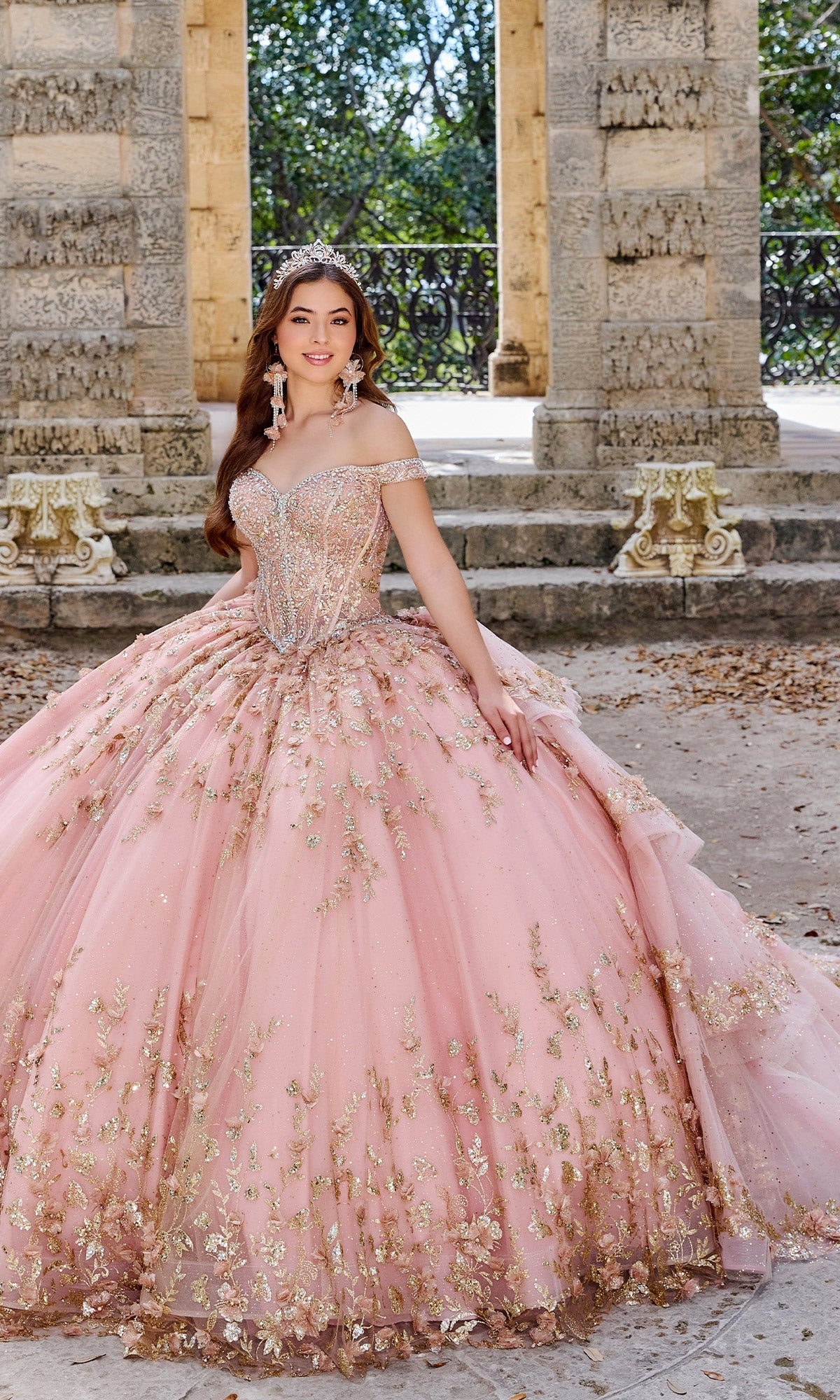 A young woman stands outdoors in front of stone columns and greenery, her Princesa PR30131 off-shoulder quinceañera dress with 3D floral appliques and pink-gold embellishments sparkling in the sunlight.